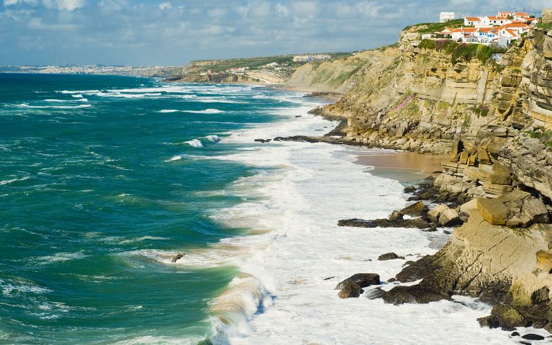 Vista panorâmica da costa de Azenhas do Mar, em Sintra, Portugal, com falésias à beira-mar, casas brancas no topo e ondas do Atlântico batendo nas rochas.