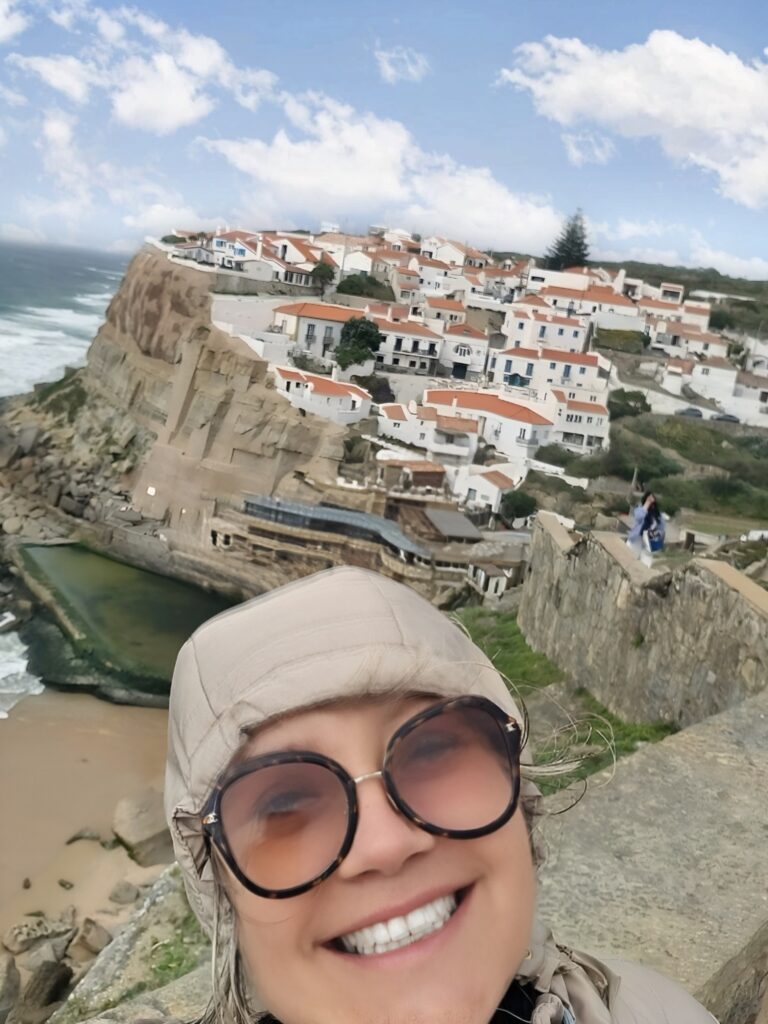 Mulher sorridente de óculos escuros faz uma selfie em frente à aldeia de Azenhas do Mar, em Portugal, com as casas brancas de telhados alaranjados erguidas sobre as falésias à beira-mar.