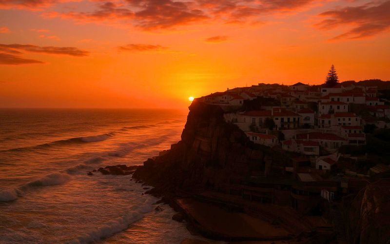 Pôr do sol em Azenhas do Mar, Portugal, com o céu em tons de laranja e vermelho iluminando as falésias e as casas brancas à beira do Atlântico.